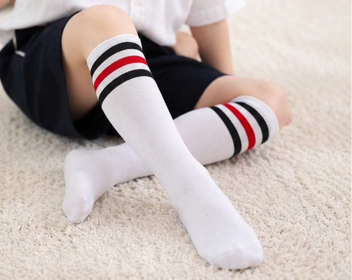 Close-up of LIUMI Kids white knee-high school socks with red and black stripe cuffs, worn by a seated child