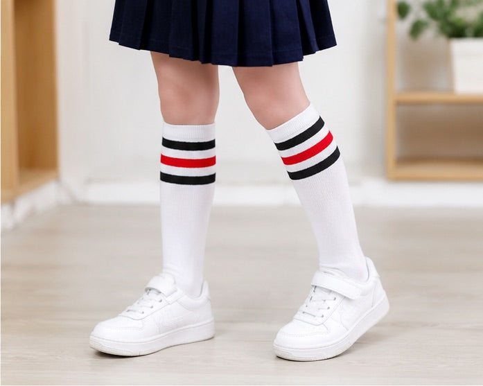 Child wearing LIUMI Kids white striped knee-high school socks with a navy pleated school skirt and white trainers, front view